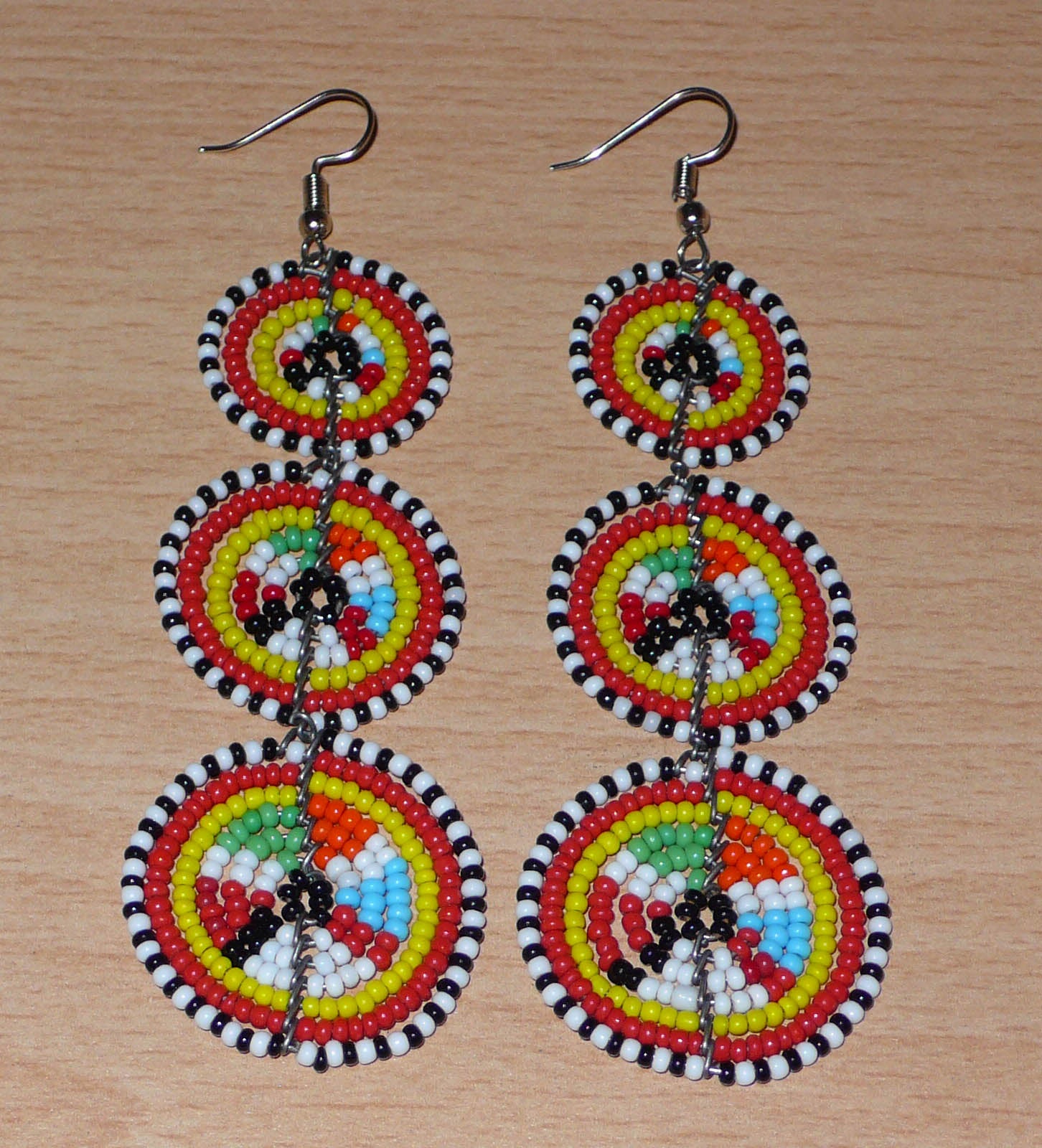 Longues boucles d'oreilles africaines de tradition Massai aux design et couleurs évoquant le style ethnique afro chic des années 70. Elles se composent de perles de rocaille multicolores glissées sur des fils de fer montés en forme de trois cercles superposés. Ces boucles d'oreilles mesurent 10.5 cm de long et 3 cm de large ; elles se portent avec des crochets en acier inoxydable sur des oreilles percées.