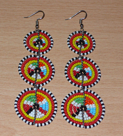 Longues boucles d'oreilles africaines de tradition Massai aux design et couleurs évoquant le style ethnique afro chic des années 70. Elles se composent de perles de rocaille multicolores glissées sur des fils de fer montés en forme de trois cercles superposés. Ces boucles d'oreilles mesurent 10.5 cm de long et 3 cm de large ; elles se portent avec des crochets en acier inoxydable sur des oreilles percées.
