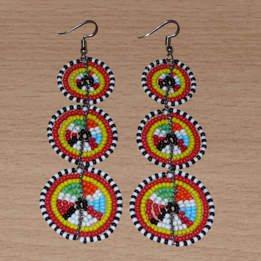 Longues boucles d'oreilles africaines de tradition Massai aux design et couleurs évoquant le style ethnique afro chic des années 70. Elles se composent de perles de rocaille multicolores glissées sur des fils de fer montés en forme de trois cercles superposés. Ces boucles d'oreilles mesurent 10.5 cm de long et 3 cm de large ; elles se portent avec des crochets en acier inoxydable sur des oreilles percées.