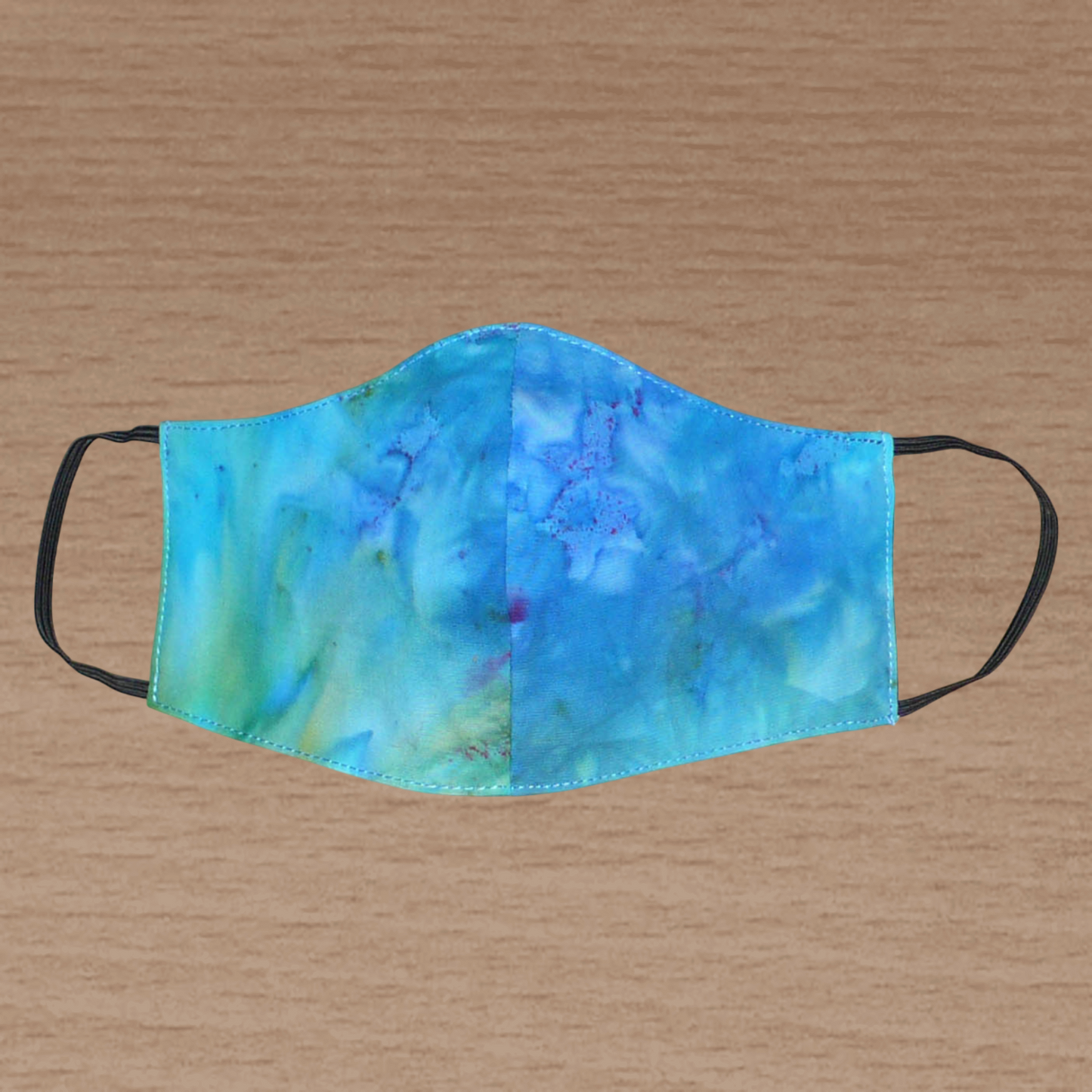 Masque facial textile à la coupe ajustée, cousu dans une cotonnade imprimée Tie and Dye bleu lagon avec des nuances de jaune et de rose ; il se porte avec des élastiques. Fabriqué en vue d'une utilisation eco-friendly, ce masque est lavable et réutilisable.