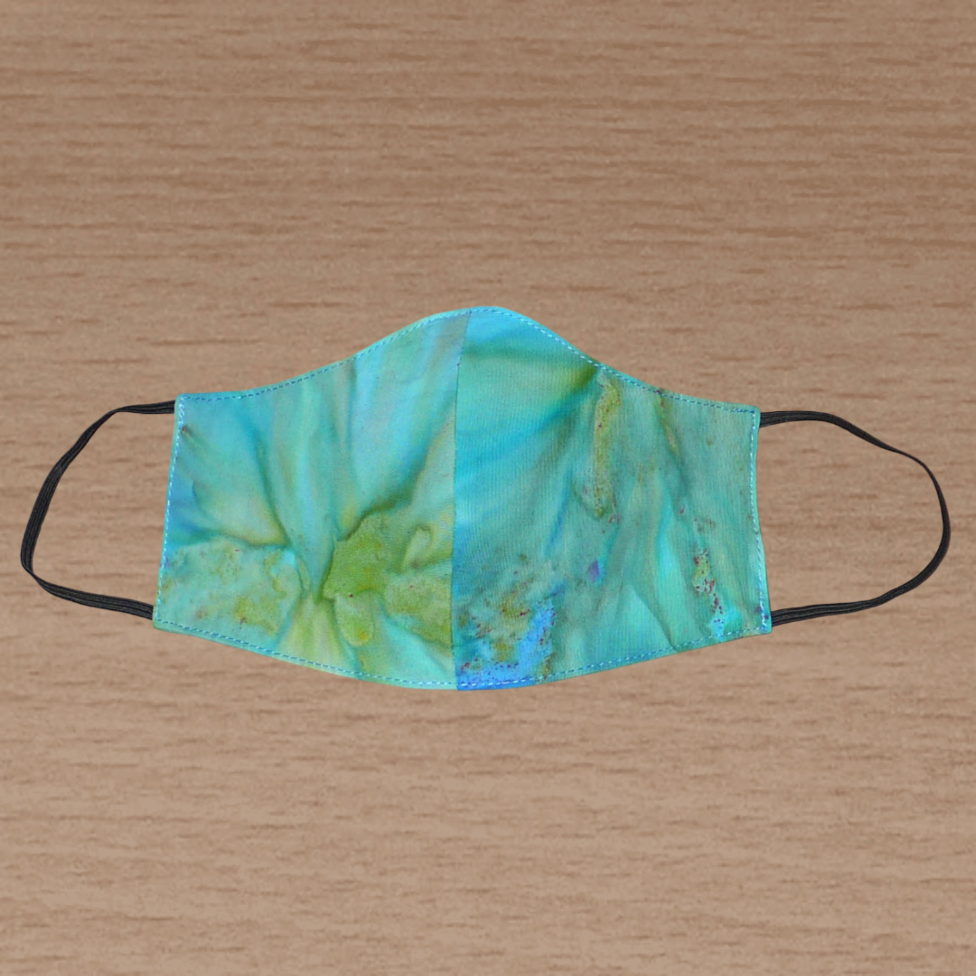 Masque facial textile en coton imprimé Tie and Dye bleu turquoise, avec des nuances de jaune et de vert ; à porter avec des élastiques.