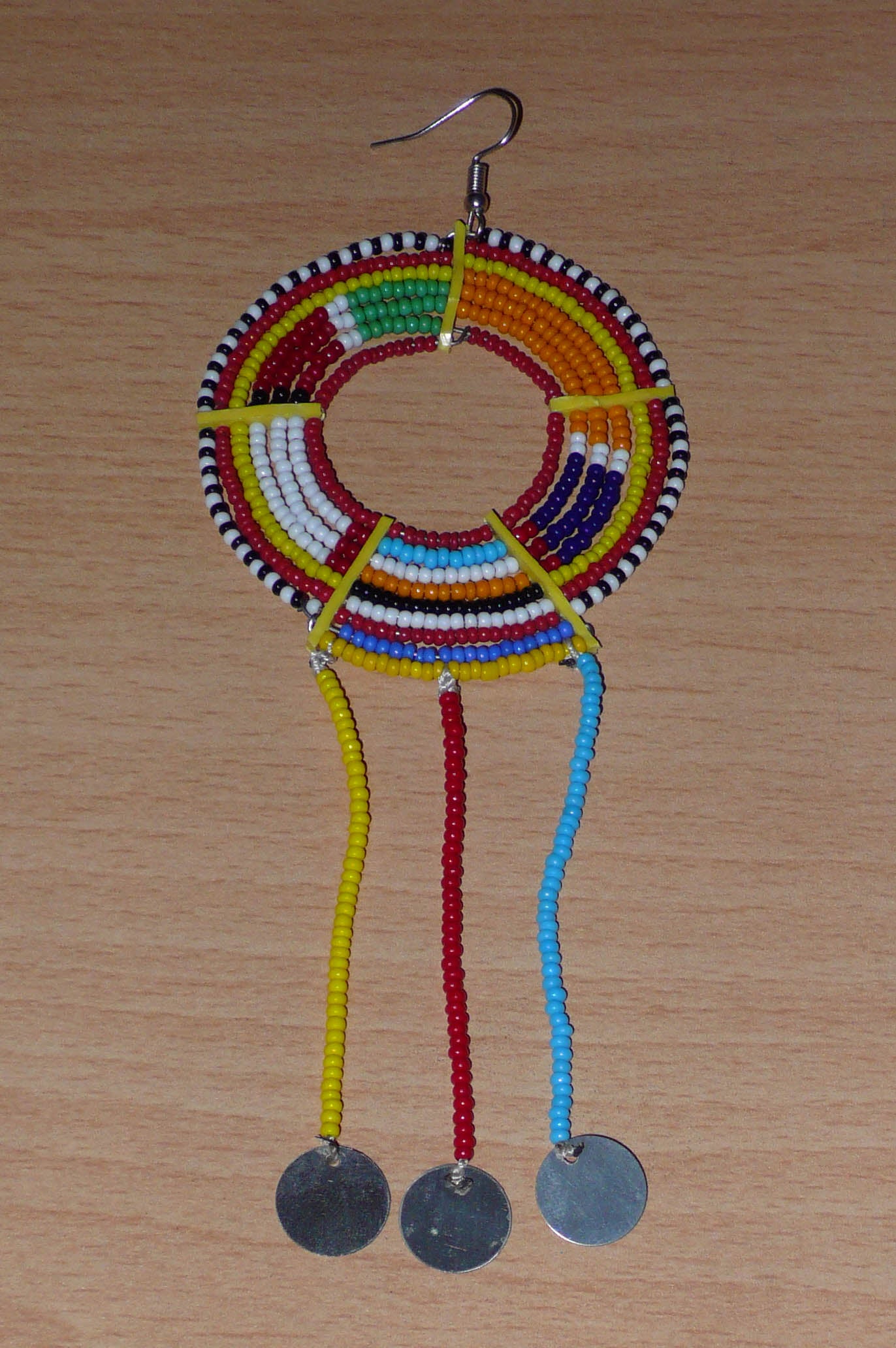 Longue mono boucle d'oreille africaine Massai à porter sur une seule oreille pour un effet visuel asymétrique. Elle se compose de perles de rocaille multicolores enfilées sur des fils de fer montés en cercles fixés sur des charnières en plastique recyclé jaune, et se prolonge de franges perlées ornées de disques en métal. Surdimensionnée, cette mono boucle d'oreille mesure 15.5 cm de long et se porte sur une oreille percée.