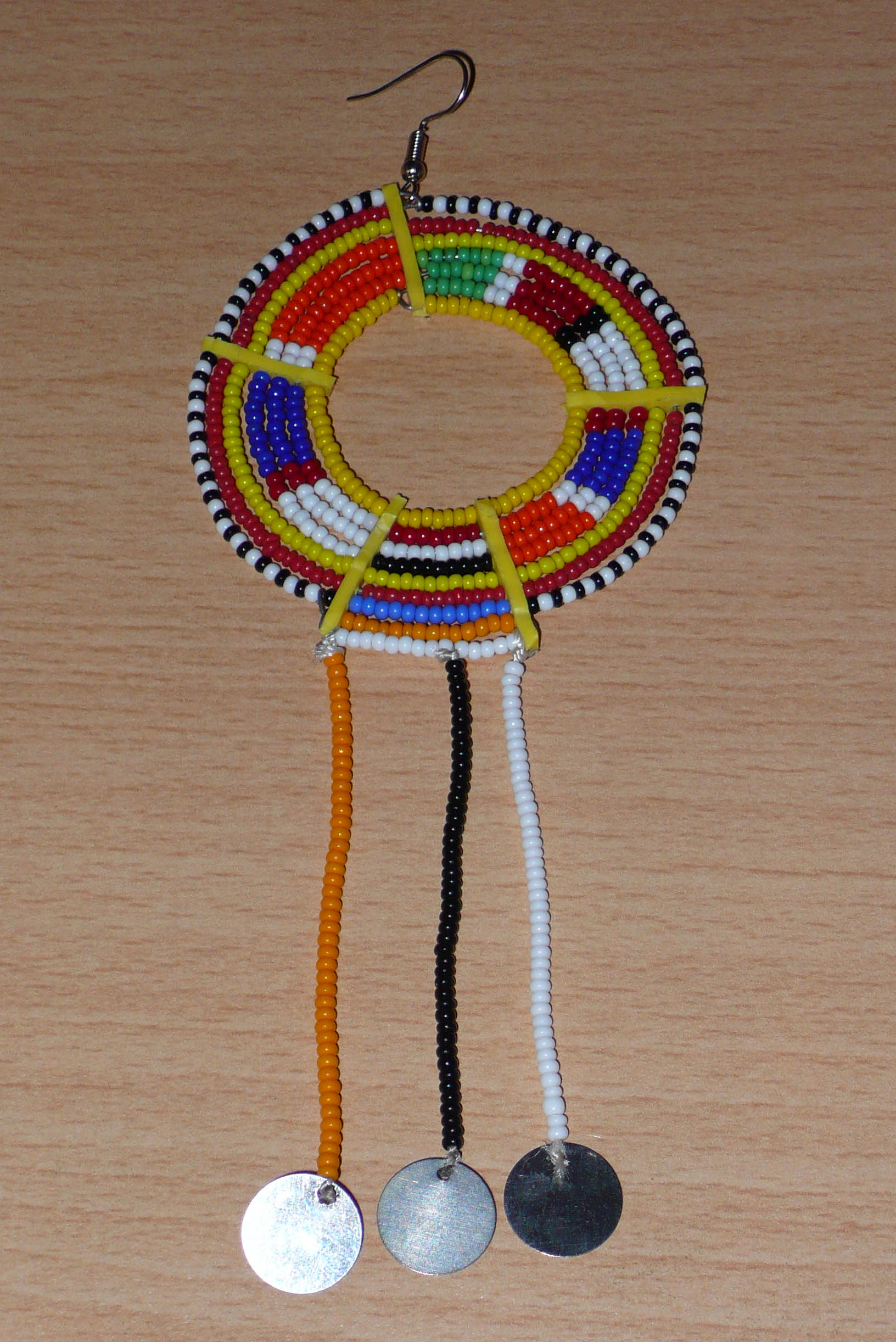 Mono boucle d'oreille confectionnée dans la plus pure tradition tribale Massai, à porter sur une seule oreille pour un effet visuel asymétrique, composée de perles de rocaille jaunes et multicolores enfilées sur des fils de fer montés en cercles et se prolonge de franges perlées ornées de disques métalliques. Surdimensionnée, elle mesure 16 cm de long et se porte sur des oreilles percées.