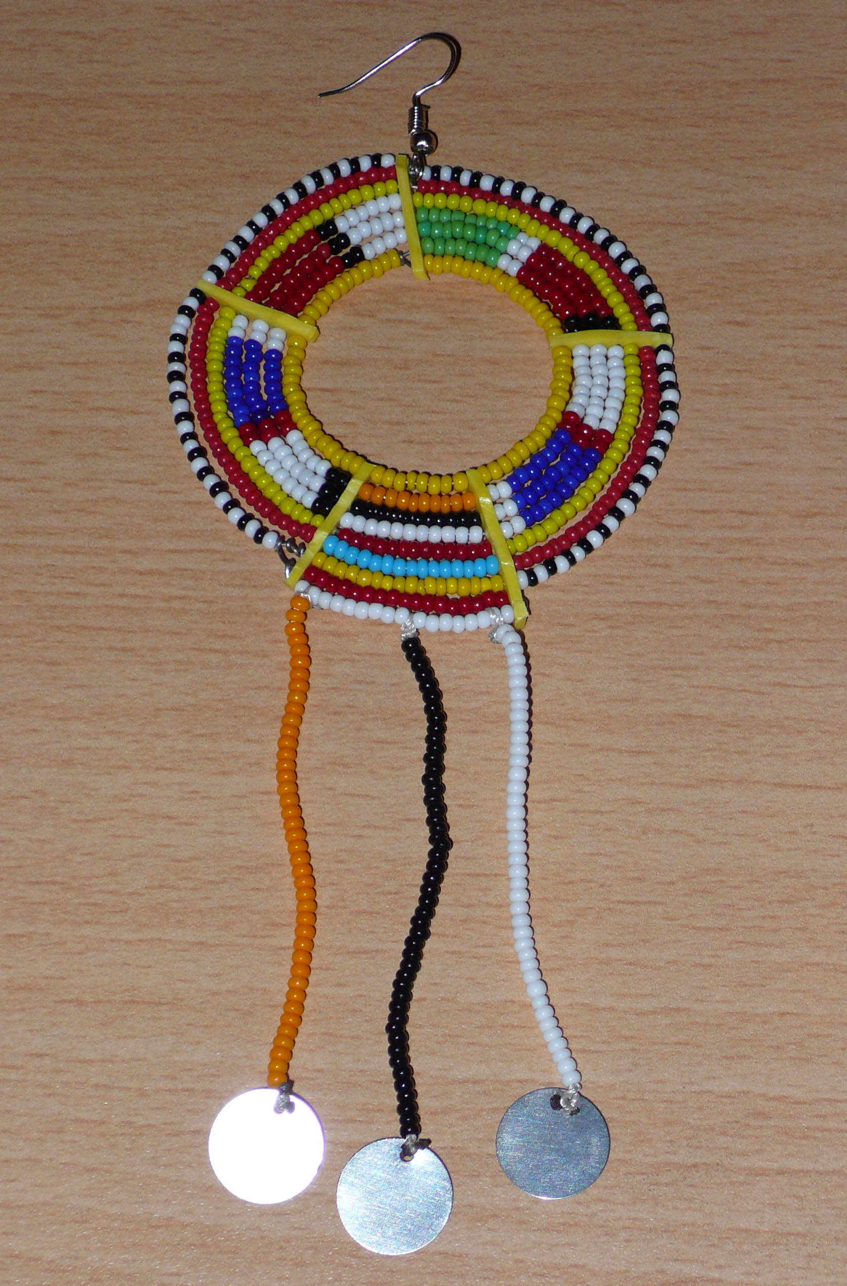 Mono boucle d'oreille à porter sur une seule oreille pour un effet visuel asymétrique : elle se compose de perles de rocaille jaunes et multicolores enfilées sur des fils de fer montés en cercles fixés sur des charnières en plastique recyclé jaune, et se prolonge de franges perlées ornées de disques métalliques. Surdimensionnée, elle mesure 16 cm de long.