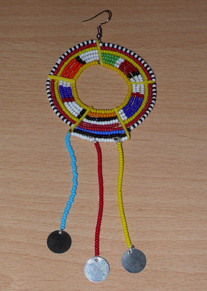 Mono boucle d'oreille à franges fabriquée au Kenya dans la tradition ethnique Massai. À porter sur une seule oreille pour un effet visuel asymétrique, ce bijou se compose de perles de rocaille multicolores glissées sur des fils de fer montés en cercles fixés sur des charnières en plastique recyclé jaune ; il se prolonge de franges perlées ornées de disques en métal. Surdimensionnée, elle mesure 16.5 cm de long et 6.5 cm de large, et se porte sur une oreille percée.