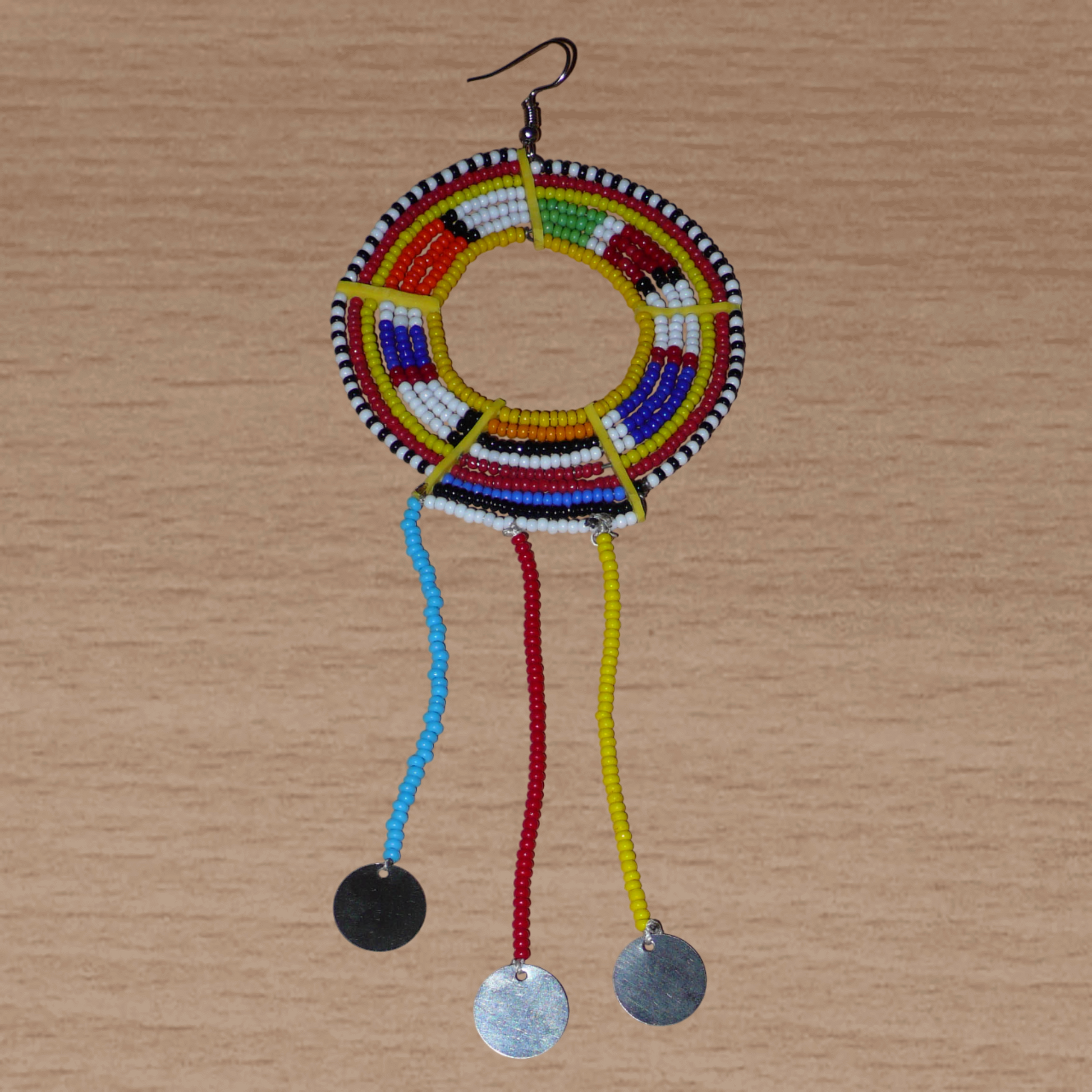 Mono boucle d'oreille à franges fabriquée au Kenya dans la tradition ethnique Massai. À porter sur une seule oreille pour un effet visuel asymétrique, ce bijou se compose de perles de rocaille multicolores glissées sur des fils de fer montés en cercles fixés sur des charnières en plastique recyclé jaune ; il se prolonge de franges perlées ornées de disques en métal. Surdimensionnée, elle mesure 16.5 cm de long et 6.5 cm de large, et se porte sur une oreille percée.