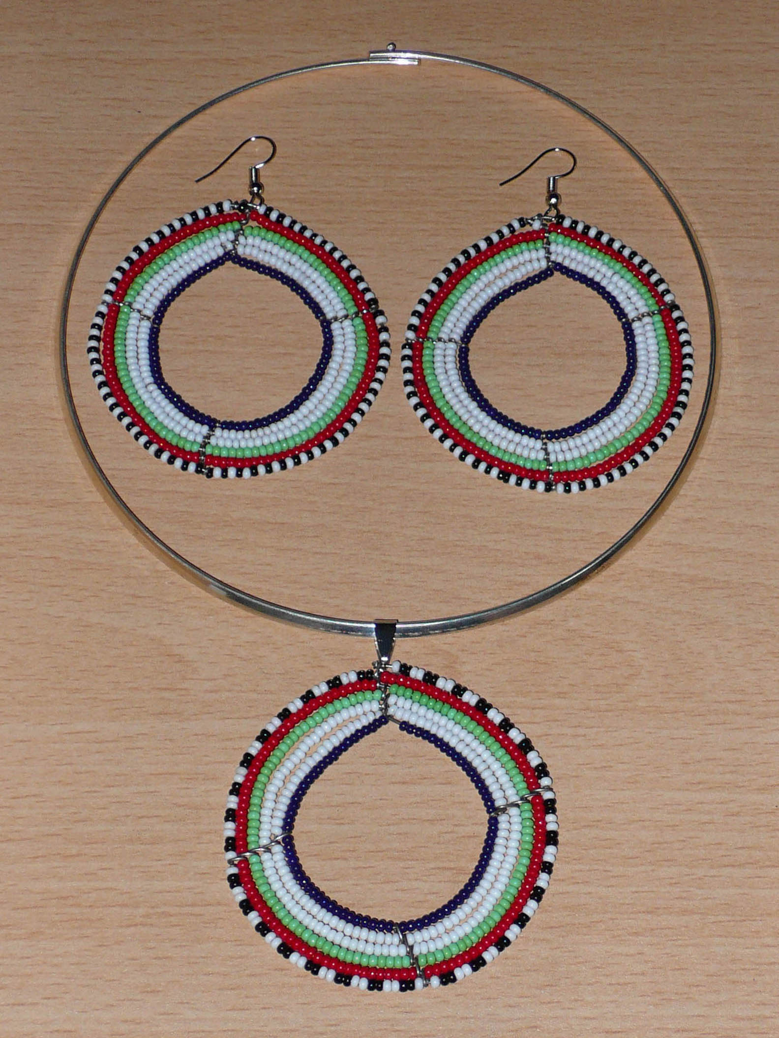 Parure de bijoux Massai au style hippie chic composée d'un collier ras-de-cou en perles de rocaille principalement blanches, puis vertes, rouges, noires et bleu marine enfilées sur des fils de fer montés en forme de cercles, ainsi que d'une paire de boucles d'oreilles rondes assorties au médaillon du collier. Timeless Fineries