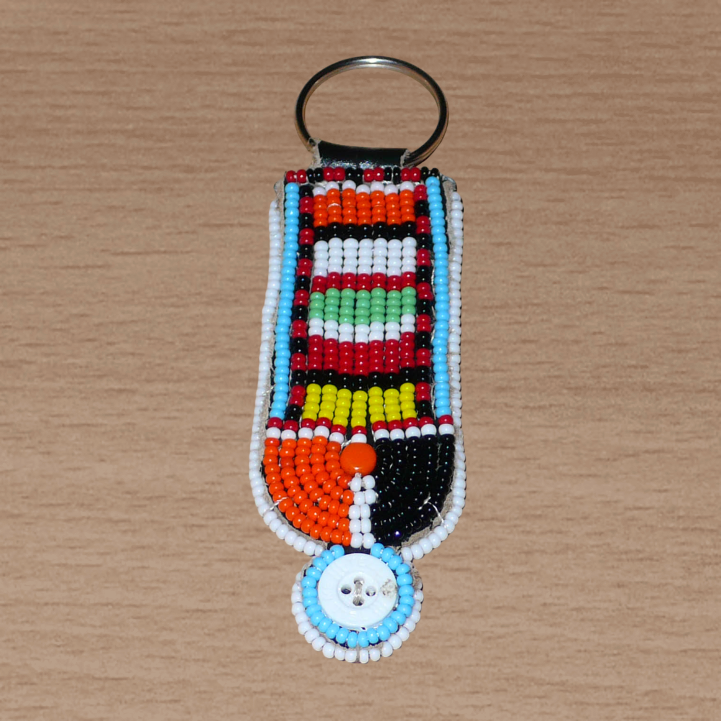 Porte-clés africain à motifs ethniques Massai composé de perles de rocaille multicolores et de boutons brodés sur un morceau de cuir. Ce porte-clés, brodé sur les deux faces, mesure 10,5 cm de long et 3 cm de large.
