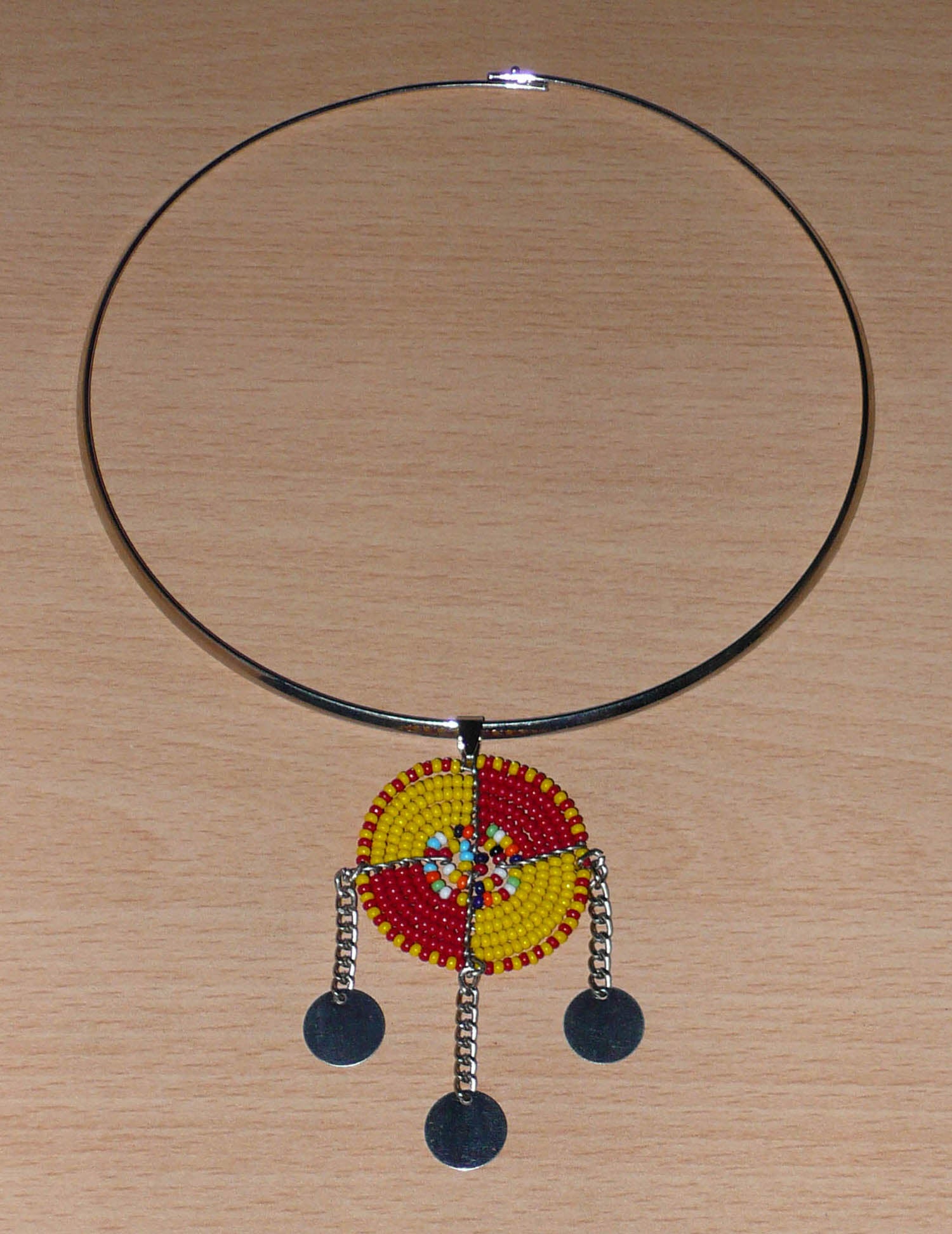 Bijou africain se composant d'un collier ras de cou en laiton rehaussé d'un pendentif Massai rond fait de perles de rocaille rouges, jaunes et multicolores, et prolongé de trois chaînettes ornées de disques métalliques.