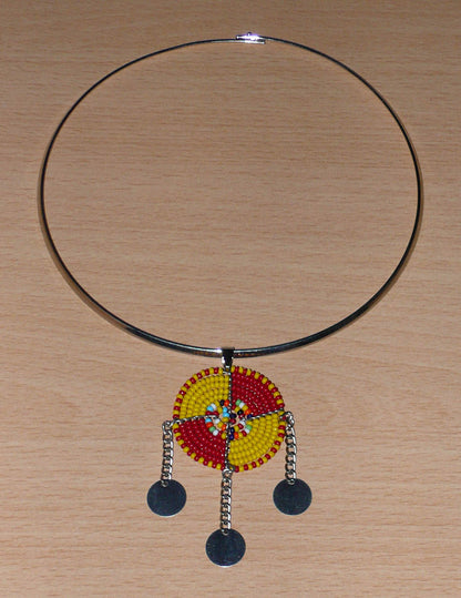 Bijou africain se composant d'un collier ras de cou en laiton rehaussé d'un pendentif Massai rond fait de perles de rocaille rouges, jaunes et multicolores, et prolongé de trois chaînettes ornées de disques métalliques.