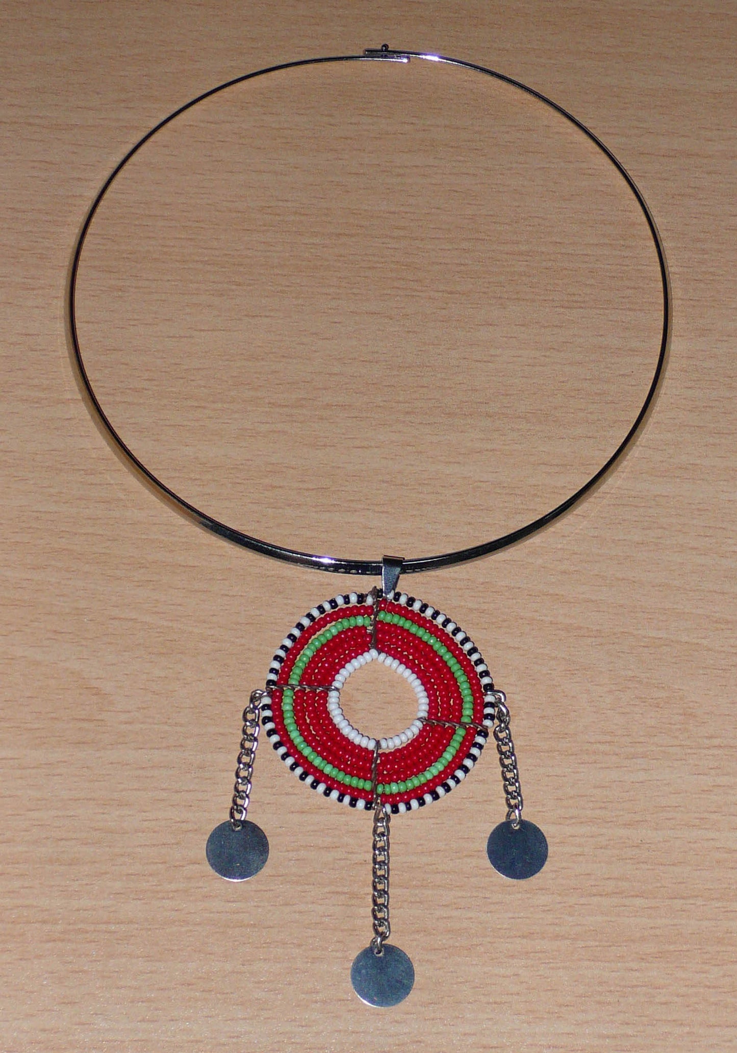 Bijou africain ethnique chic se composant d'un ras de cou en laiton agrémenté d'un beau pendentif Massai de forme ronde fait de perles de rocaille principalement rouges et vertes, et prolongé de trois chaînettes décorées de disques métalliques.