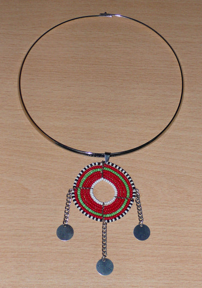 Bijou africain ethnique chic se composant d'un ras de cou en laiton agrémenté d'un beau pendentif Massai de forme ronde fait de perles de rocaille principalement rouges et vertes, et prolongé de trois chaînettes décorées de disques métalliques.
