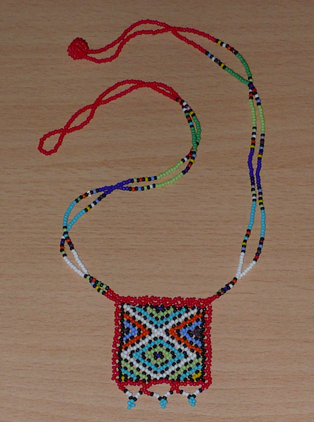 Collier africain composé de perles de rocaille rouges agrémenté d'un pendentif carré aux motifs multicolores zoulous. Il mesure 27,5 cm en partant de la nuque jusqu'à la base du pendentif. Le pendentif mesure 5,5 cm sur 4,5 cm. Le collier s'attache avec un fermoir à boule. Timeless Fineries
