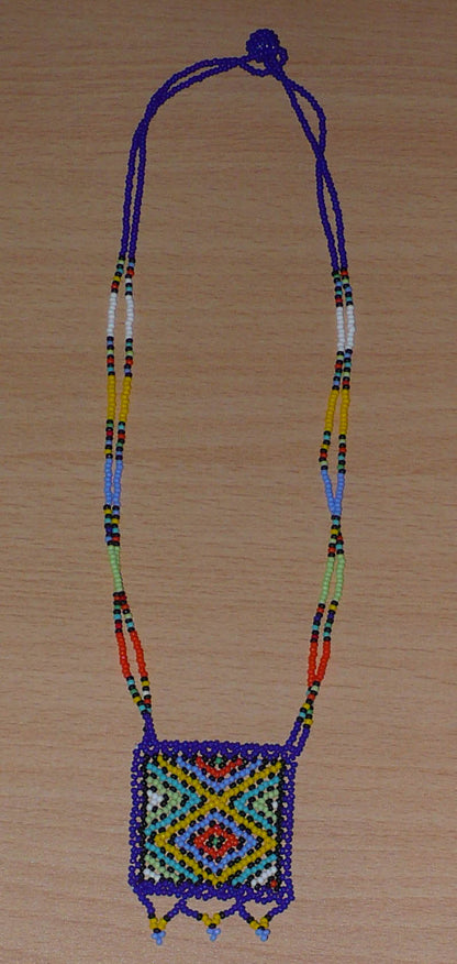 Collier africain en perles de rocaille, agrémenté d'un pendentif carré orné de motifs traditionnels zoulous bleus et multicolores. Il mesure 27,5 cm de long en partant de la nuque jusqu'à la base du pendentif. Le pendentif mesure 5,5 cm sur 4,5 cm. Le collier s'attache avec un fermoir à boule. Timeless Fineries