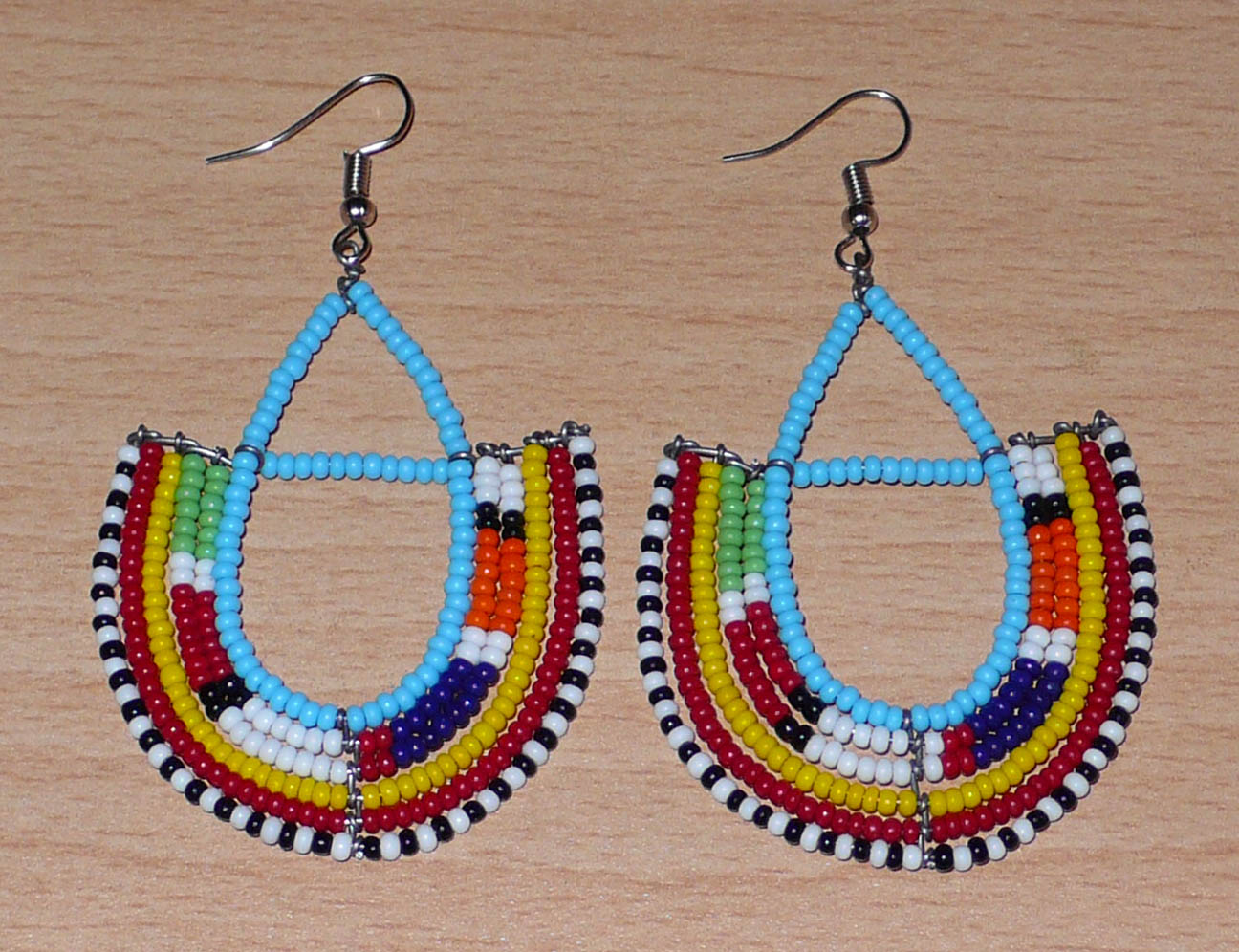 Boucles d'oreilles africaines à motifs ethniques Massai fabriquées au Kenya. Elles se composent de perles de rocaille bleu ciel et multicolores montées en forme d'ancres. Ces boucles d'oreilles afro chic mesurent 7,5 cm de long et 4,5 cm de large. Elles se portent avec des crochets en acier inoxydable sur des oreilles percées.