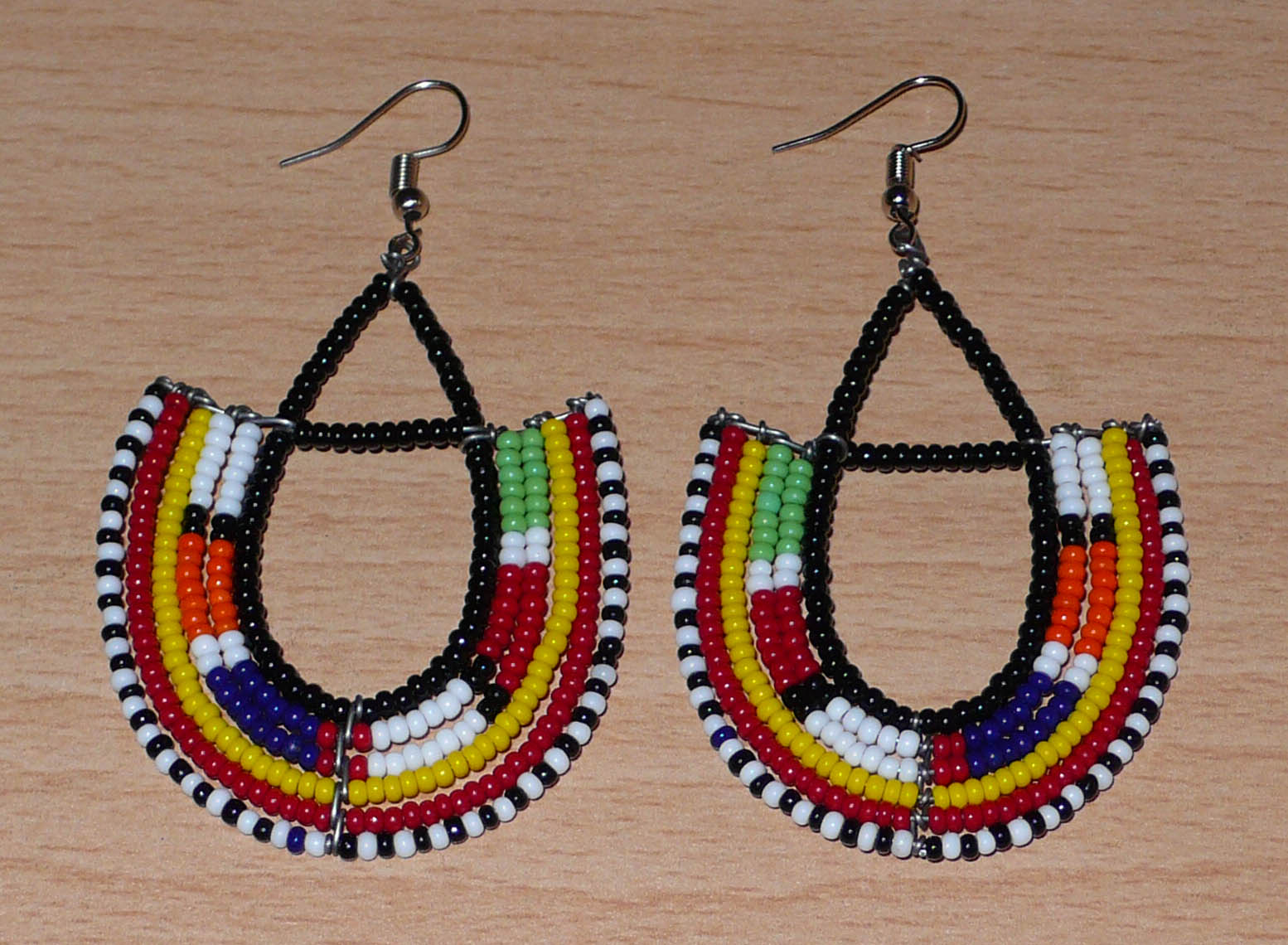 Boucles d'oreilles africaines traditionnelles fabriquées au Kenya par des femmes de la tribu Massai ; elles se composent de perles de rocaille noires et multicolores montées en forme d'ancres, et mesurent 7,5 cm de long et 4,5 cm de large. Elles se portent sur des oreilles percées avec des crochets en acier inoxydable.