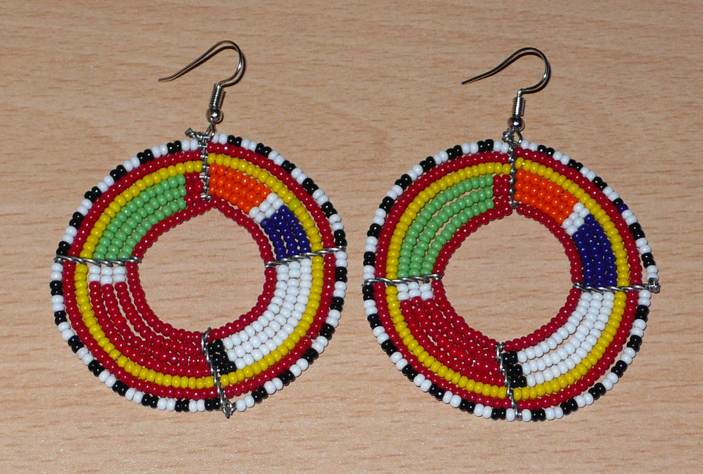 Boucles d'oreilles africaines rondes aux motifs et couleurs Massai composées de perles de rocaille rouges et multicolores enfilées sur sept rangées de fils de fer montés en forme de cercles. Elles mesurent 7,5 cm de long et 5,5 cm de large et se portent avec des crochets en acier inoxydable sur des oreilles percées.