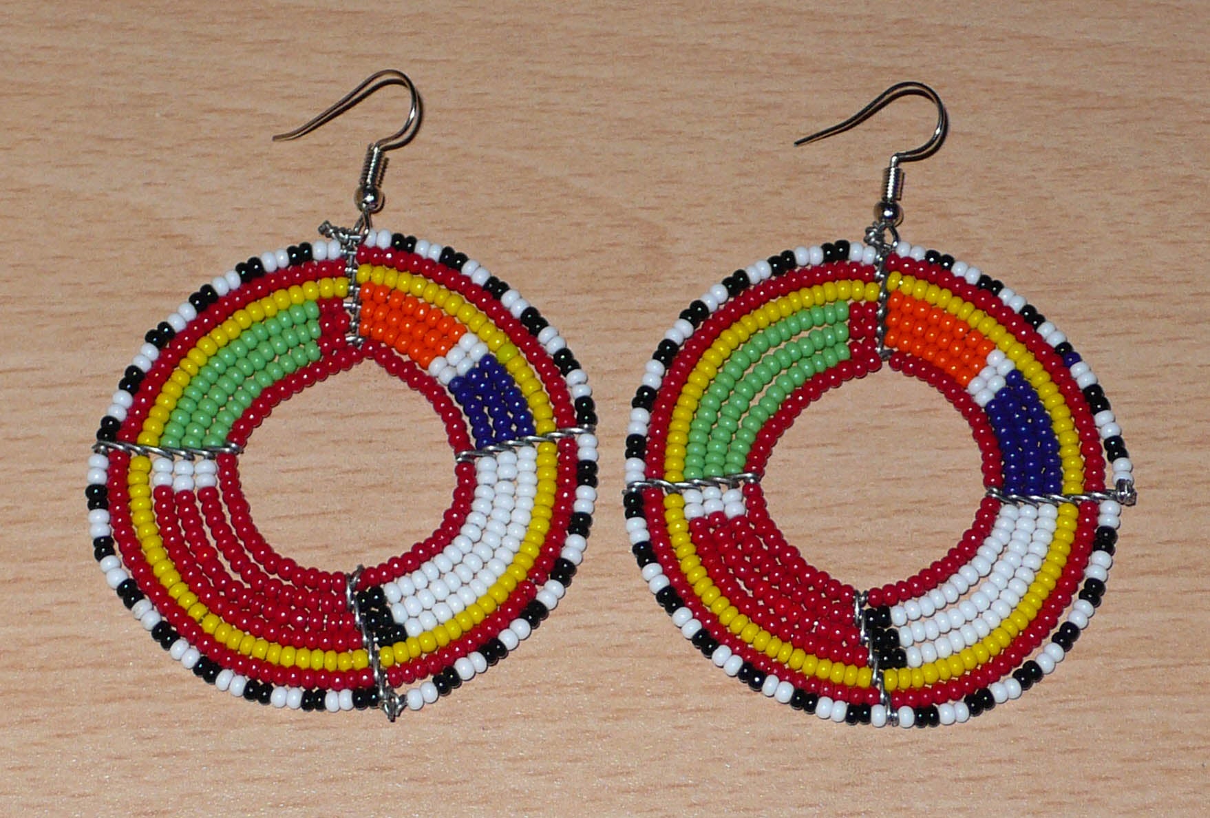 Boucles d'oreilles africaines rondes aux motifs et couleurs Massai composées de perles de rocaille rouges et multicolores enfilées sur sept rangées de fils de fer montés en forme de cercles. Elles mesurent 7,5 cm de long et 5,5 cm de large et se portent avec des crochets en acier inoxydable sur des oreilles percées.