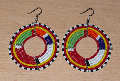 Boucles d'oreilles africaines rondes aux motifs et couleurs Massai composées de perles de rocaille rouges et multicolores enfilées sur sept rangées de fils de fer montés en forme de cercles. Elles mesurent 7,5 cm de long et 5,5 cm de large et se portent avec des crochets en acier inoxydable sur des oreilles percées.