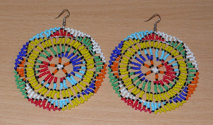 Boucles d'oreilles sud-africaines rondes au style afro chic des années 70. Elles se composent de perles de rocaille multicolores enfilées sur des fils de nylon fixés sur des structures circulaires. Surdimensionnées, elles mesurent 10 cm de long et 8,5 cm de large, et se portent sur des oreilles percées.