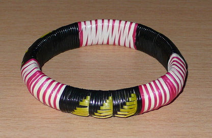 Bracelet africain eco-friendly fabriqué au Mali ; il se compose de motifs tressés avec du plastique recyclé noir, jaune, rose et blanc.  Ce bracelet a une circonférence de 19,5 cm, un diamètre de 6 cm et une épaisseur de 1,5 cm.  Il peut être porté par une femme ou par un homme. Timeless Fineries