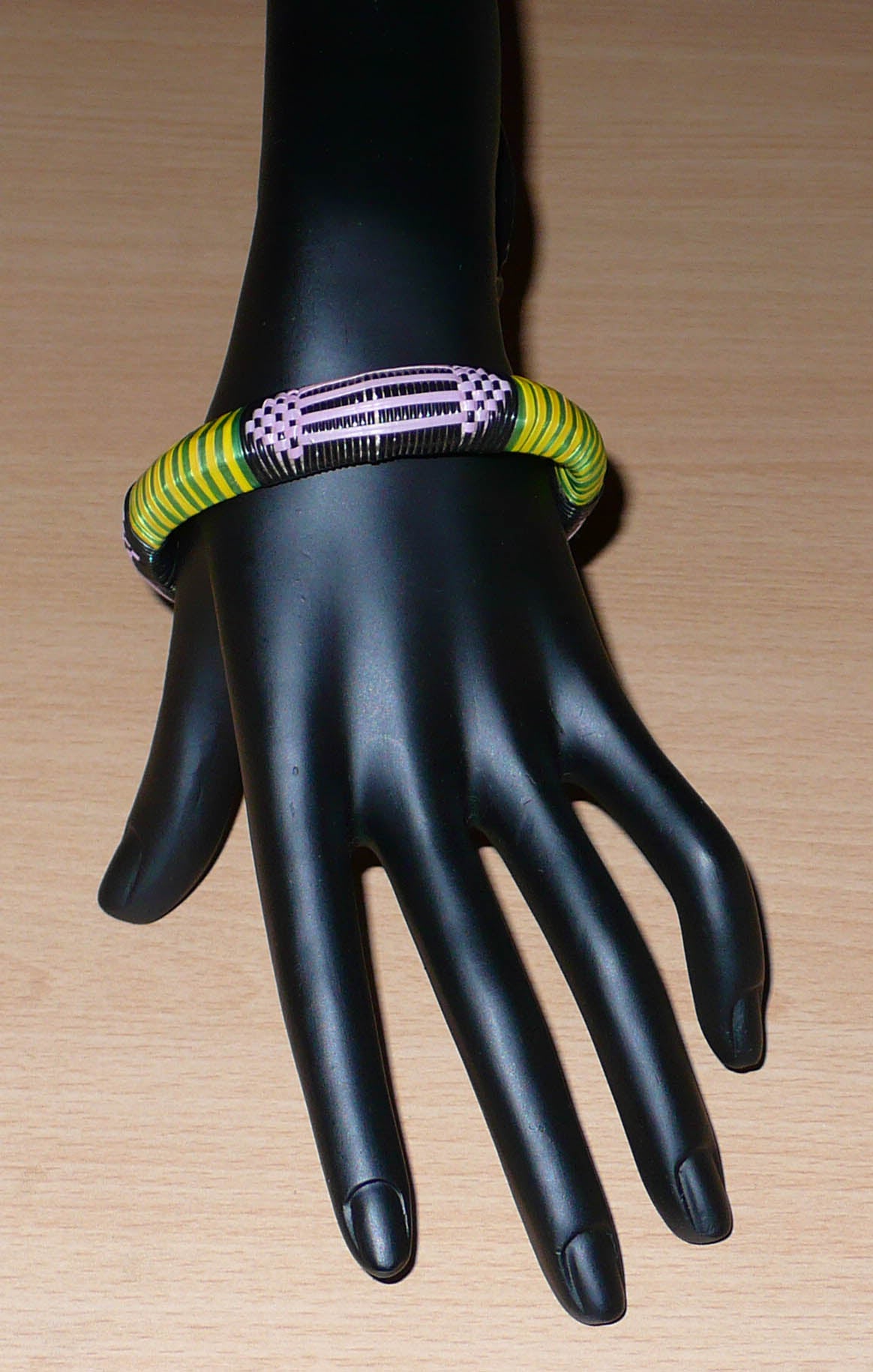 Fabriqué dans une démarche éco-responsable, ce bracelet africain à motifs tribaux est tissé avec de fines bandes de plastique recyclé noir, parme, vert et jaune.  La circonférence interne du bracelet est de 20,5 cm, son diamètre de 6,5 cm et son épaisseur de 1,3 cm.  Il peut être porté par une femme ou par un homme.