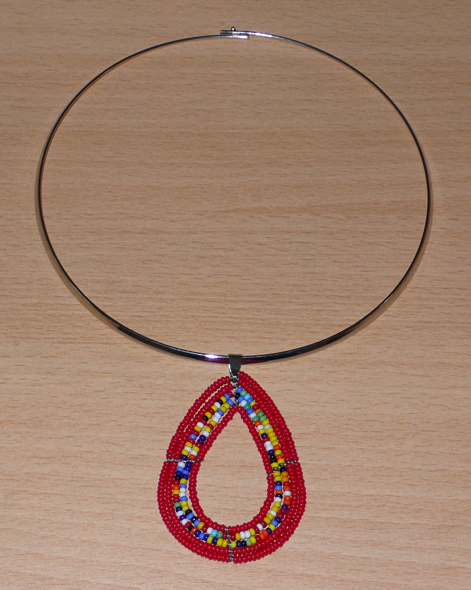 Collier ethnique de style afro bohème composé d'un ras de cou en laiton orné d'un pendentif en forme de goutte fait de perles de rocaille rouges et multicolores. Il mesure 20 cm de long, la circonférence du ras de cou en laiton est de 42,5 cm et son diamètre de 13,5 cm ; le pendentif perlé mesure 7 cm de long et 4,5 cm de large.
