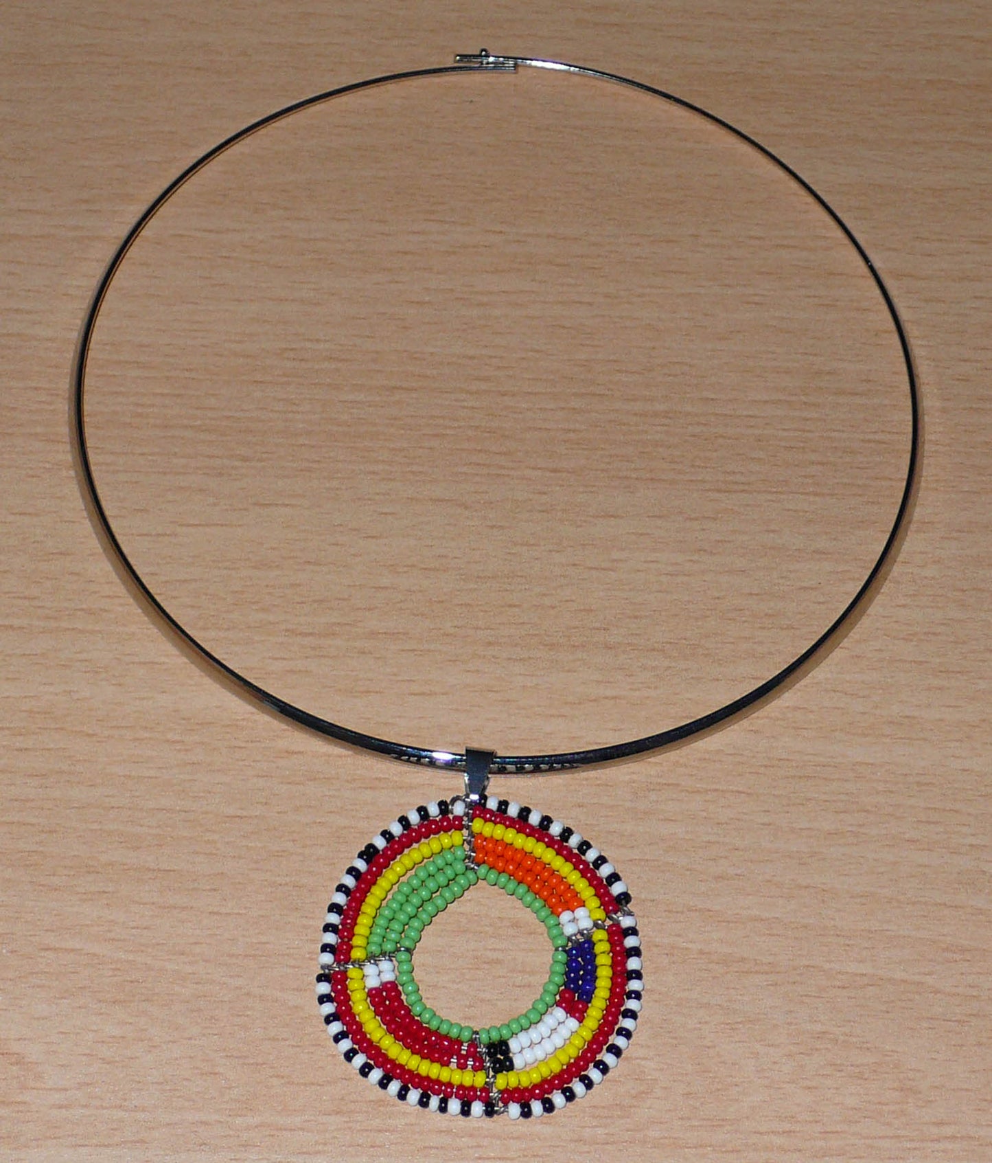 Collier africain composé d'un ras de cou en laiton argenté orné d'un médaillon Massai en perles de rocaille vertes et multicolores. Le collier mesure 19 cm de long (de la nuque jusqu'à la base du pendentif), la circonférence du tour de cou en laiton est de 42,5 cm avec un diamètre de 13,5 cm ; le médaillon mesure 5 cm de long et de large. 