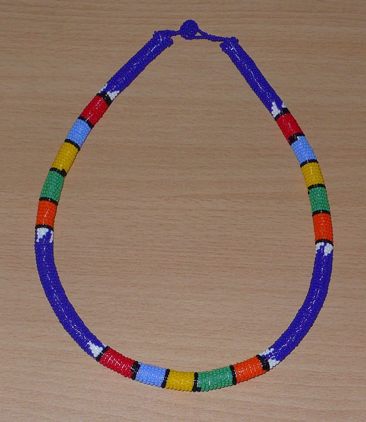 Bijou ethnique bleu. Collier sud-africain tubulaire bleu et multicolore. Il se compose d'un tube de plastique autour duquel sont enroulées des petites perles de rocaille bleues alternées avec des motifs ethniques traditionnels zoulous rouges, bleu ciel, jaunes, verts et orange. Le collier mesure 22 cm de long (de la nuque jusqu'à la base du collier), la longueur du tour de cou du collier est 56 cm, et son épaisseur est de 1 cm. Il s'attache avec un fermoir à boule. Timeless Fineries