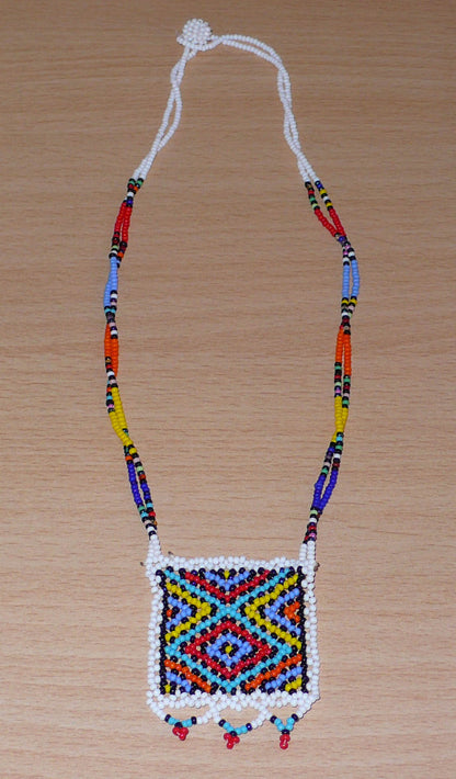 Collier africain en perles de rocaille blanches et multicolores agrémenté d'un pendentif carré à motifs ethniques. Il mesure 27 cm de long (en partant de la nuque jusqu'à la base du pendentif), le pendentif mesure 5,5 cm sur 4,5 cm ; le collier s'attache avec un fermoir à boule. Timeless Fineries