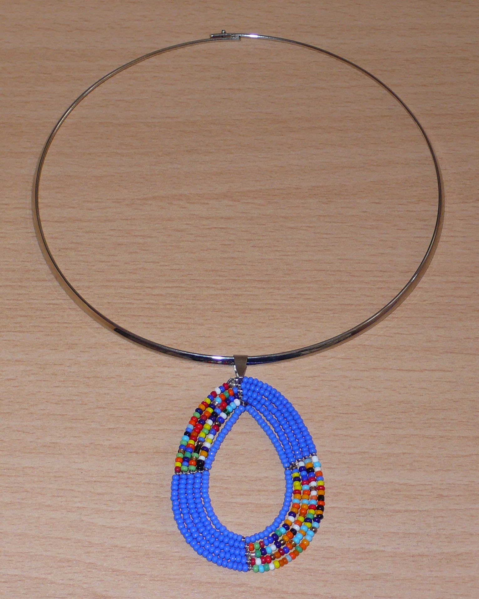 Collier bleu composé d'un ras-de-cou en laiton argenté orné d'un pendentif fait de perles de rocaille bleues et multicolores enfilées sur cinq rangs de fils de fer montés en forme de goutte. Ce collier mesure 19,5 cm de long (de la nuque au bas du pendentif), la cironférence du ras-de-cou est de 42,5 cm et 13,5 cm de diamètre ; le pendentif mesure 6 cm de long et 4,5 cm de large. Timeless Fineries