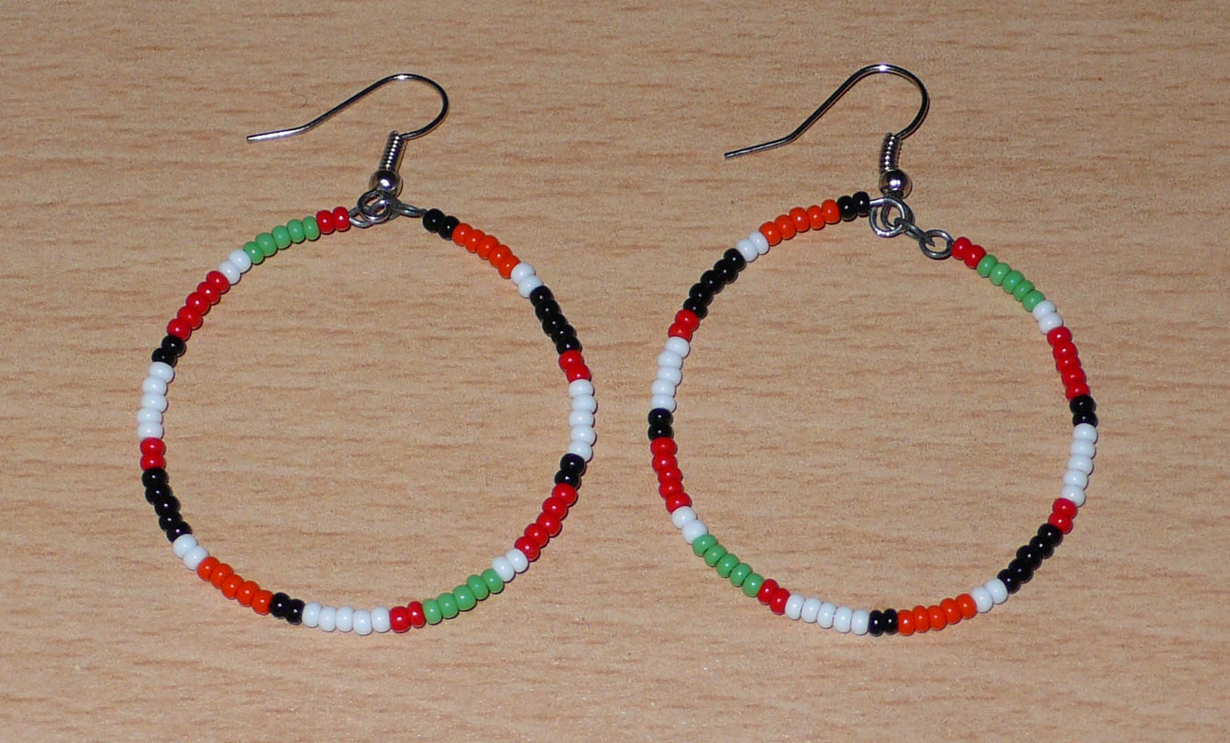 Créoles aux couleurs ethniques Massai faites de perles de rocaille rouges, vertes, noires, blanches et orange glissées sur un brin de fils de fer monté en cercle. Elles mesurent 6 cm de long et 4 cm de large, et se portent avec des crochets en acier inoxydable sur des oreilles percées.