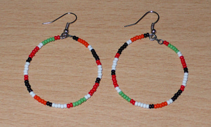Créoles aux couleurs ethniques Massai faites de perles de rocaille rouges, vertes, noires, blanches et orange glissées sur un brin de fils de fer monté en cercle. Elles mesurent 6 cm de long et 4 cm de large, et se portent avec des crochets en acier inoxydable sur des oreilles percées.