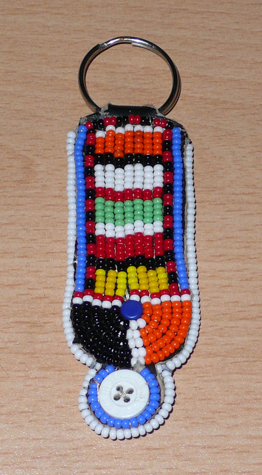 Porte-clés africain à motifs traditionnels Massai composé de perles de rocaille multicolores et de boutons brodés sur un morceau de cuir. Ce porte-clés, brodé sur les deux faces, mesure 10,5 cm de long et 3 cm de large.