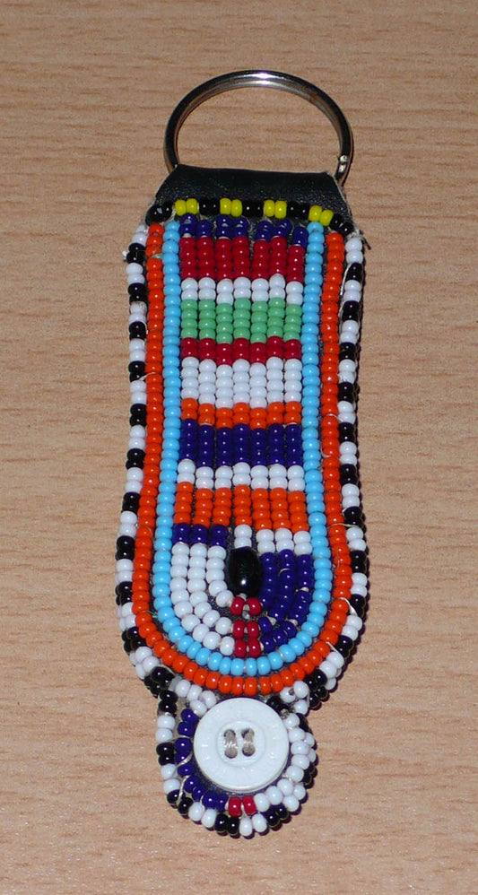Porte-clés africain à motifs traditionnels Massai composé de perles de rocaille multicolores et de boutons brodés sur un morceau de cuir. Ce porte-clés, brodé sur les deux faces, mesure 10,5 cm de long et 3 cm de large. Timeless Fineries